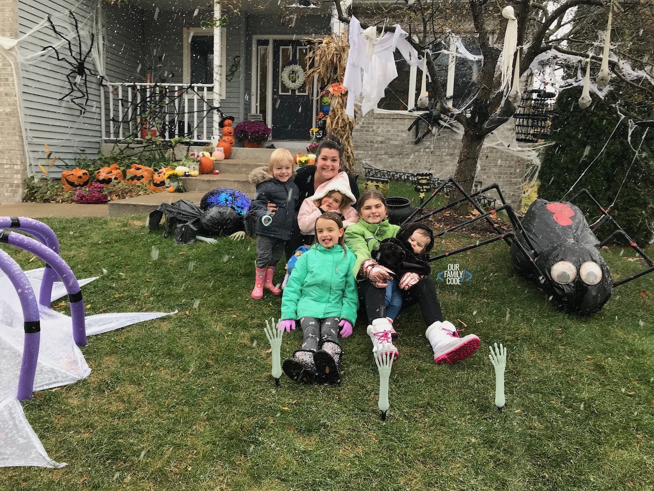 A picture of kids and mom in front of decorated house for Halloween with giant PVC spiders and hanging ghosts.
