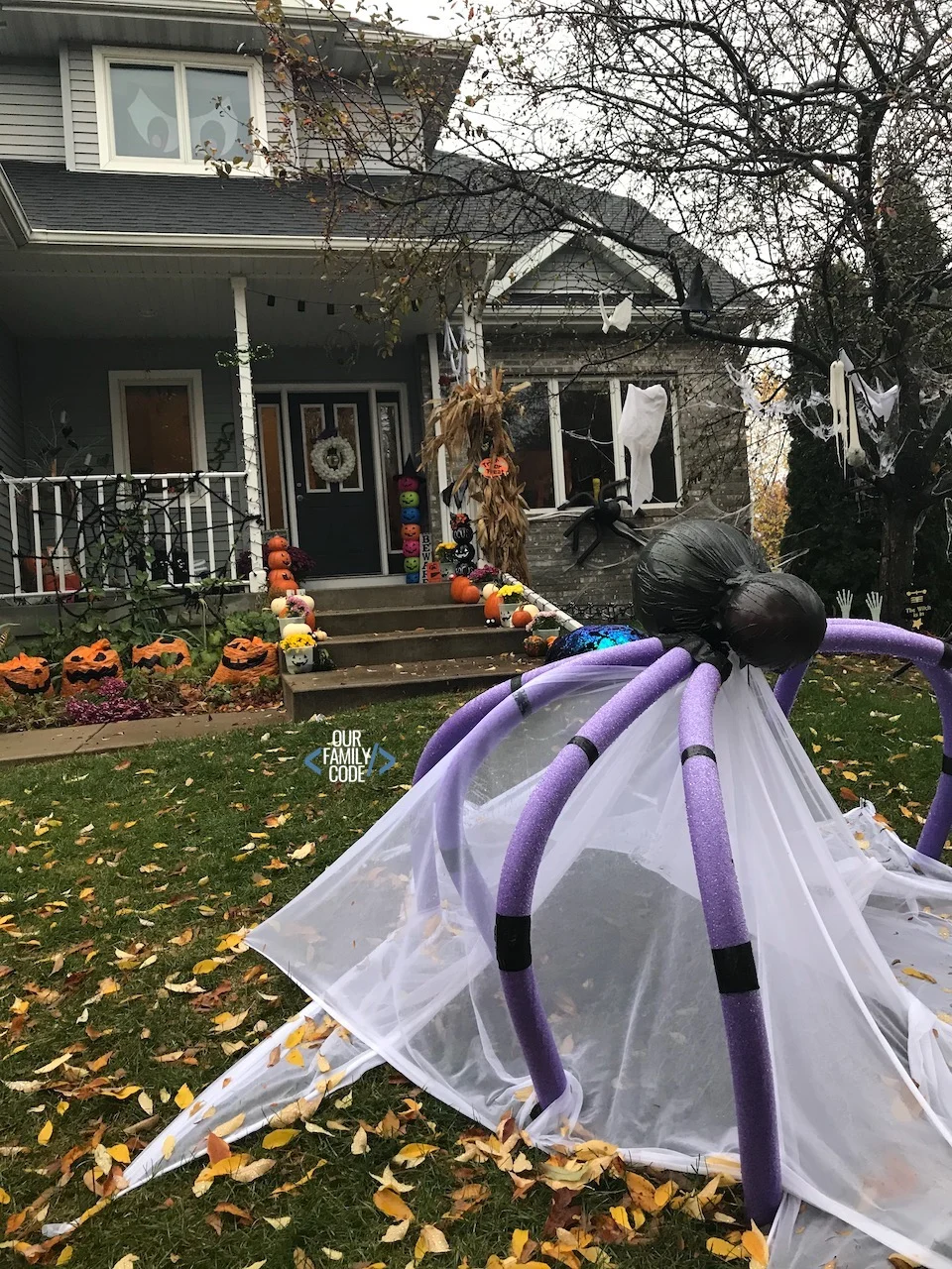 A picture of a house decorated for Halloween with a plastic pumpkin tower decoration, lots of pumpkins, hanging ghosts, and a giant pool noodle spider.