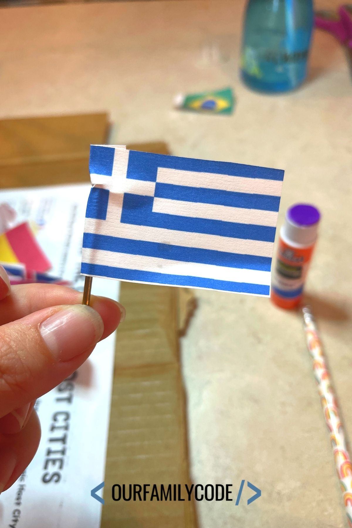 A picture of the national flag of Greece with a small nail used to mark the location of the country on a paper map.